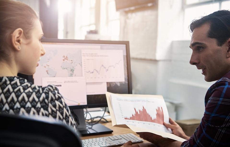 Two investors sit at a desk in front of a computer while one explains a printout of a chart to the other.