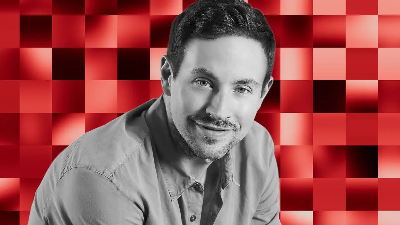 Professional portrait of a man with short hair against a red and pink checkered background.
